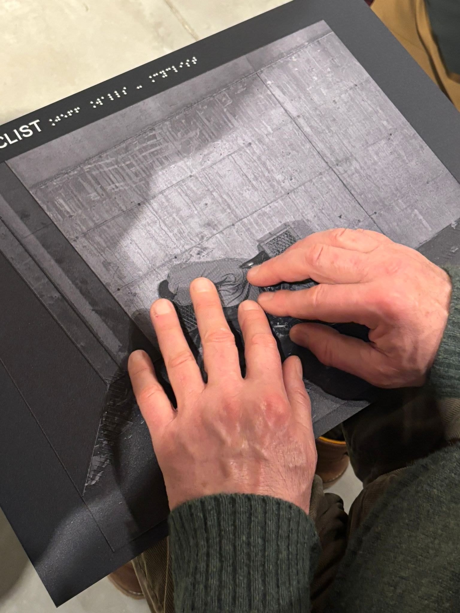Due mani esplorano il rilievo di un pannello tattile che riproduce una fotografia di Jeff Wall; in alto il titolo in lettere e in braille