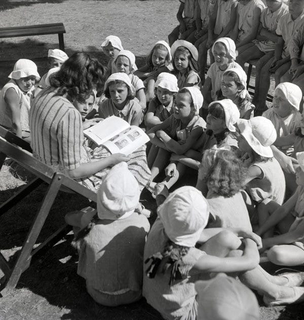 Una donna legge un libro a un gruppo di bambine con cappellini bianchi