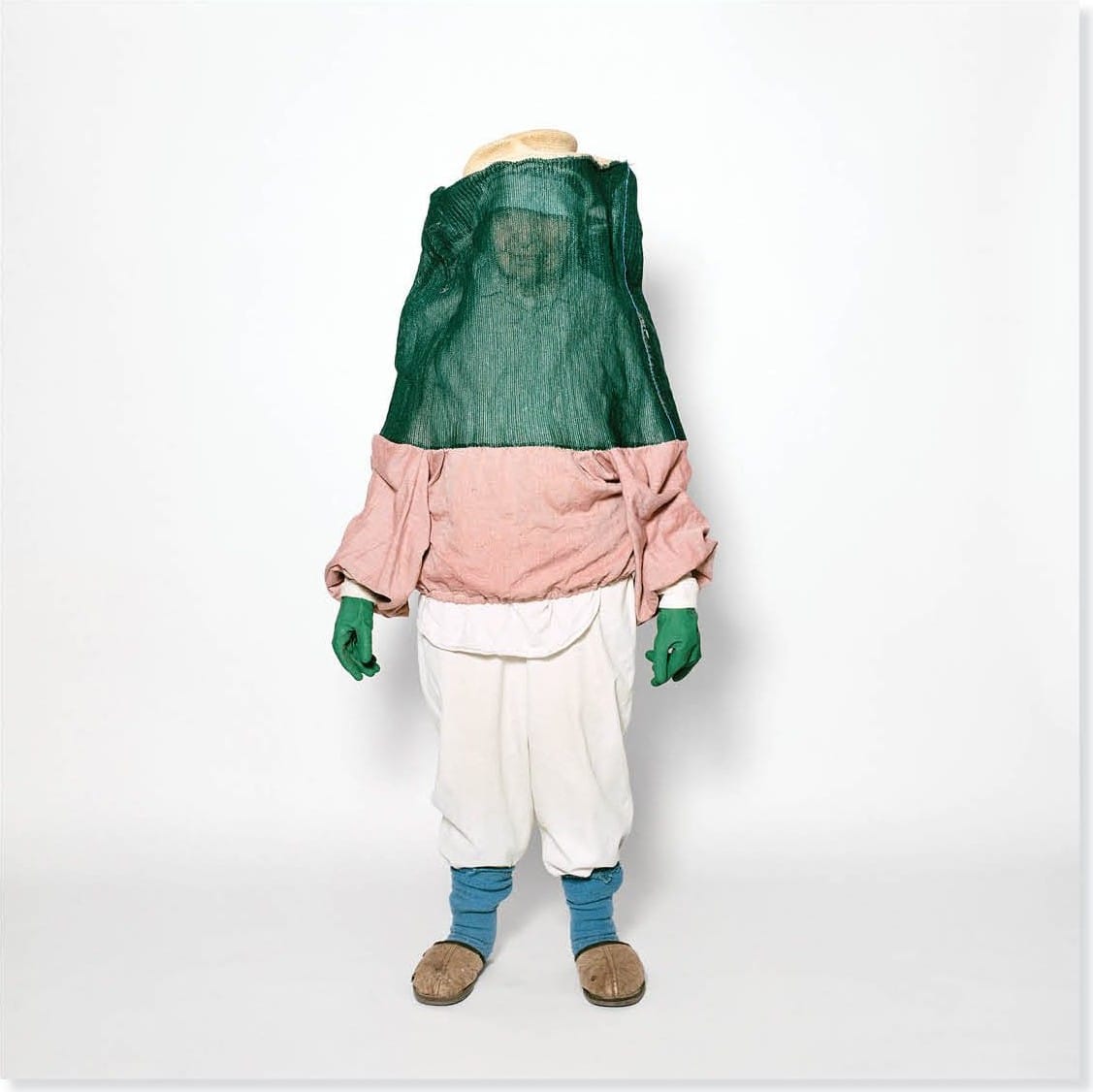 Fotografia di Kai Wasikowski: una figura con il volto coperto da una rete verde, camicia rosa e guanti verdi