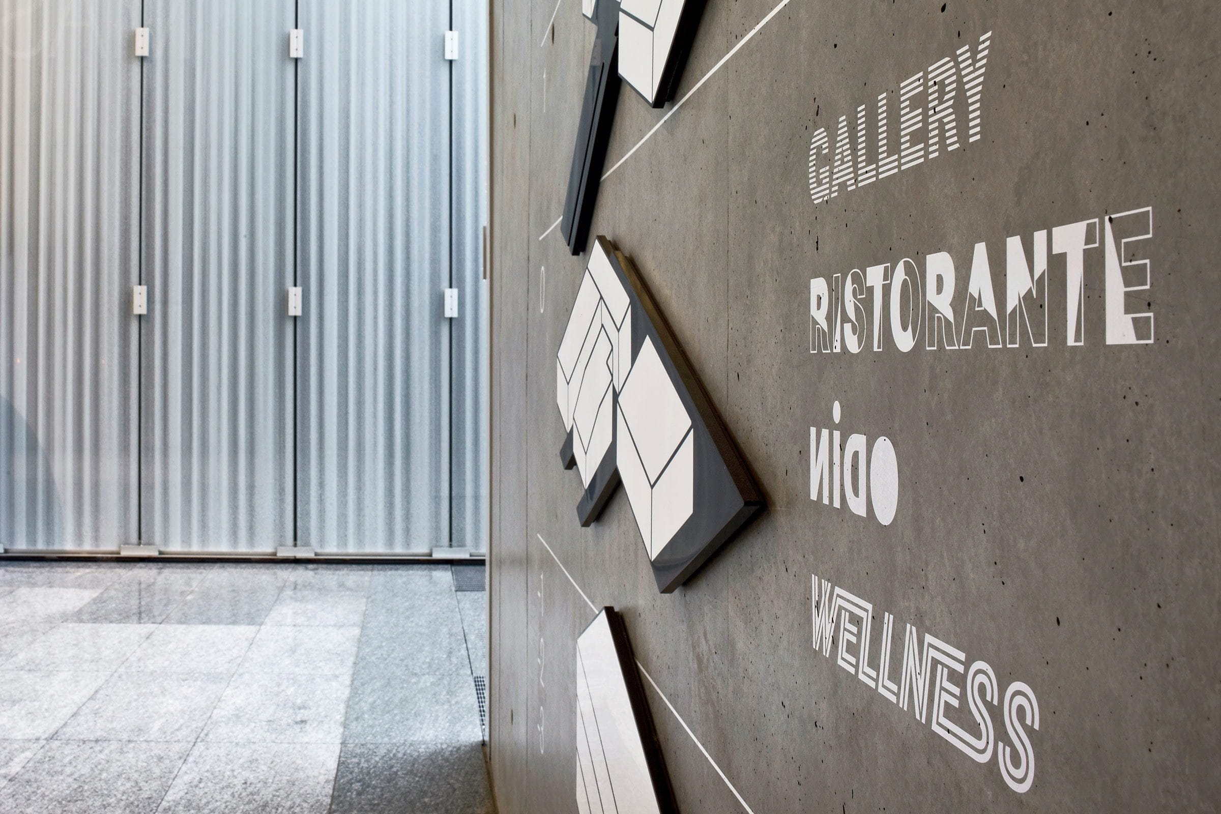 La parola WELLNESS incisa sul muro di cemento del livello 0, sotto Gallery, ristorante e Nido