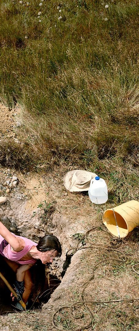 Bambina accovacciata accanto a una buca scavata nell’erba, con una tanica e un secchio
